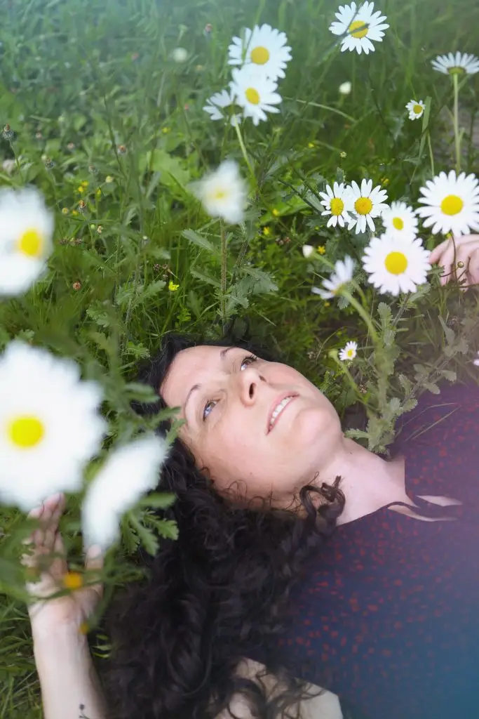 Femme allongée dans un champ de marguerites, regard paisible tourné vers le ciel, incarnant la reconnexion à soi, la détente et l’harmonie avec la nature