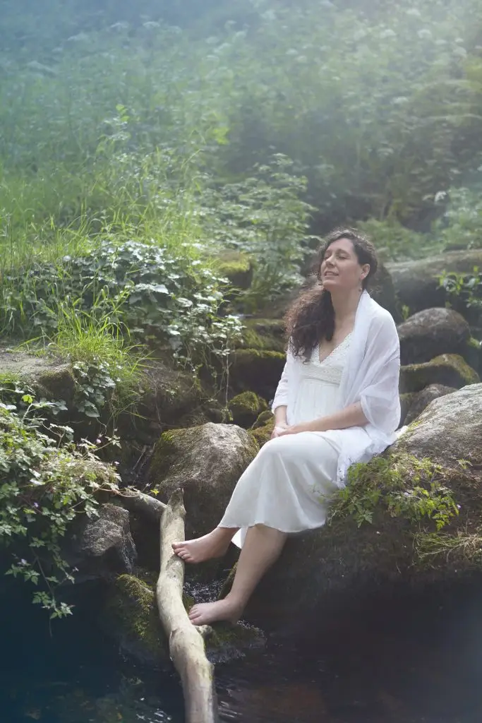 Femme en robe blanche assise sur un rocher dans la forêt, Elle tend son visage vers la lumière. Elle s'aime et se sent sereine.